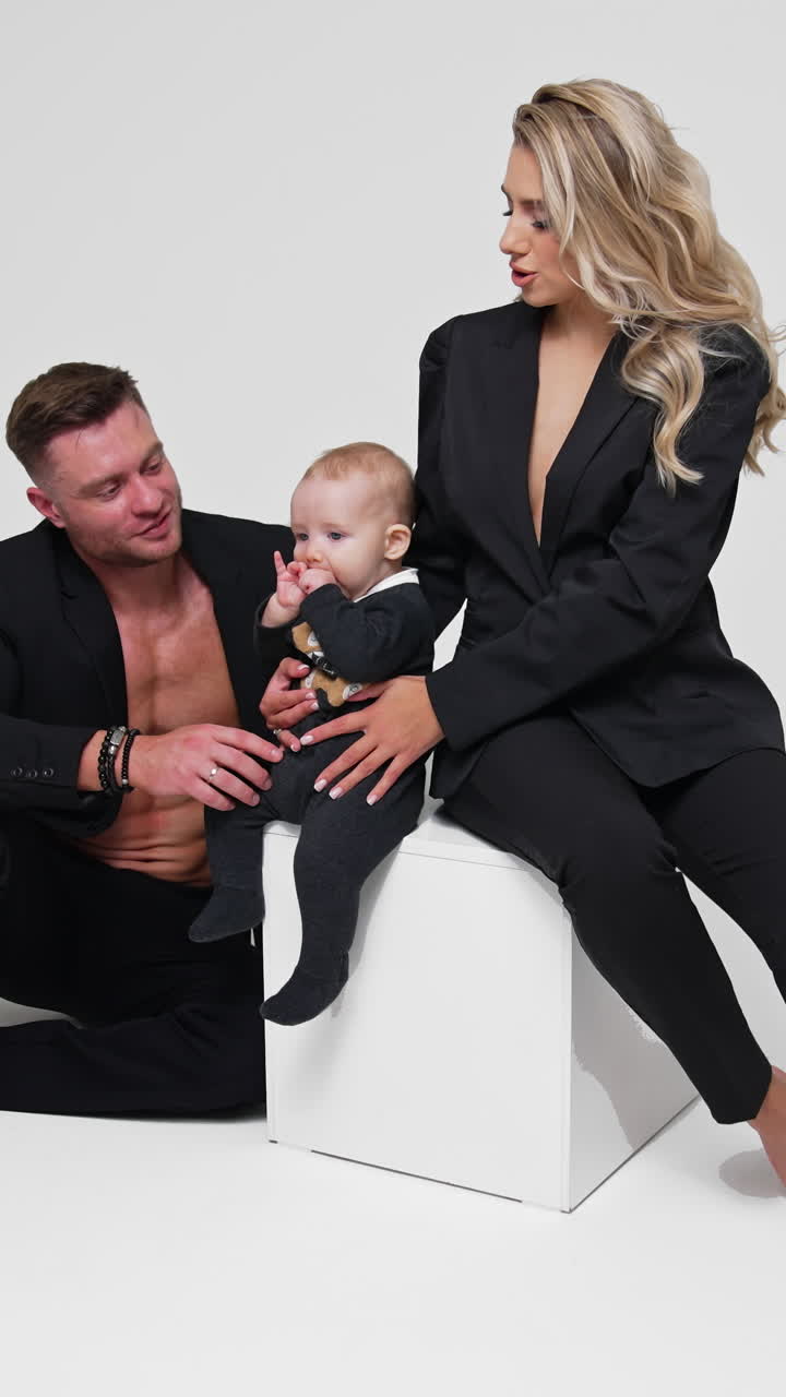 Hermosa mujer rubia se sienta en un cubo blanco sosteniendo a su bebé. Un hombre se sienta junto al cubo jugando con el niño. Grabación de estudio. Fondo blanco. Video vertical.