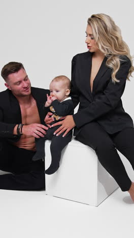 beautiful blonde woman sits on white cube holding her baby. man sits beside the cube playing with child. studio footage. white backdrop. vertical video