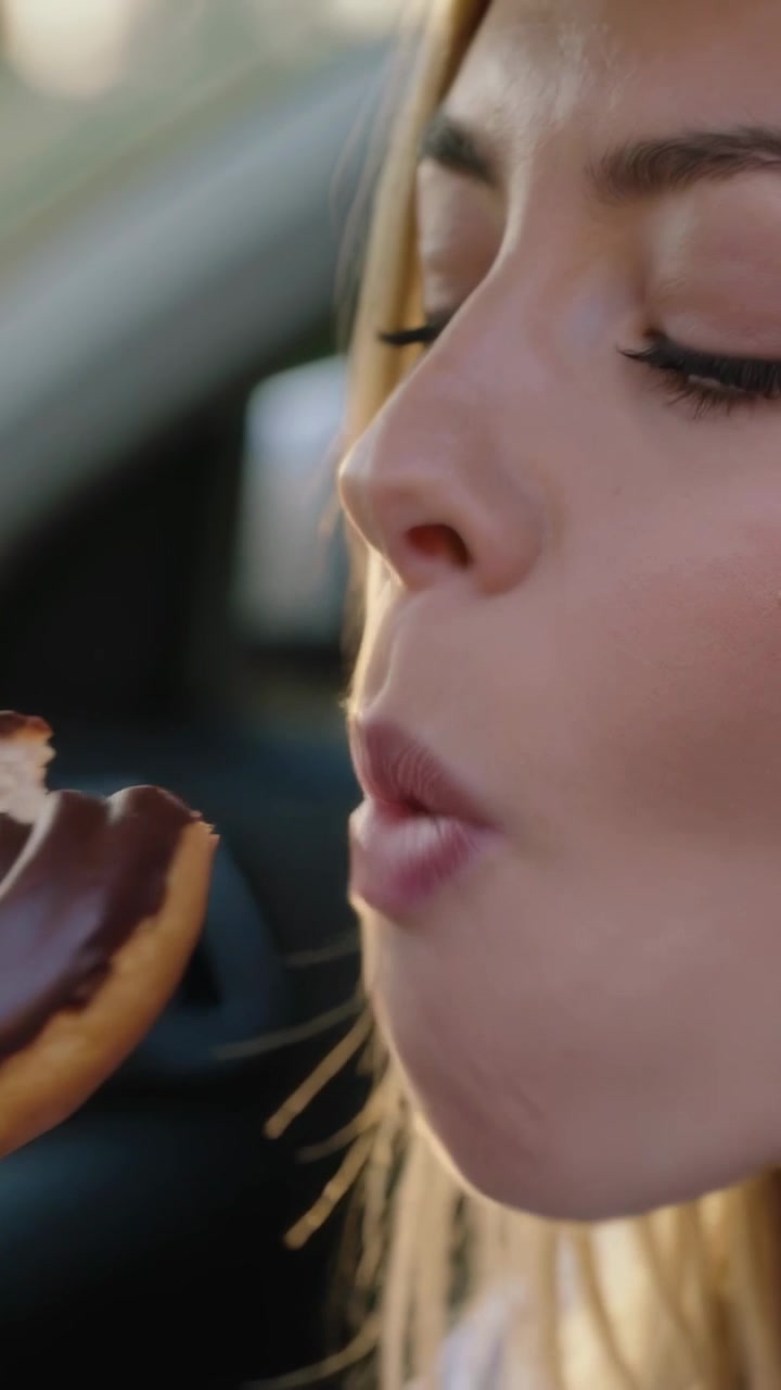 Vertical video: Taking bite, woman biting chocolate-glazed donut in parked car, holding right hand