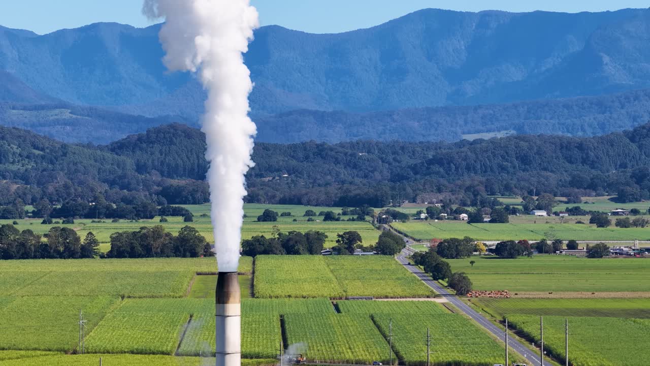 Aerial footage captures steam rising from a sugarcane factory amidst lush fields and distant mountains under clear blue skies