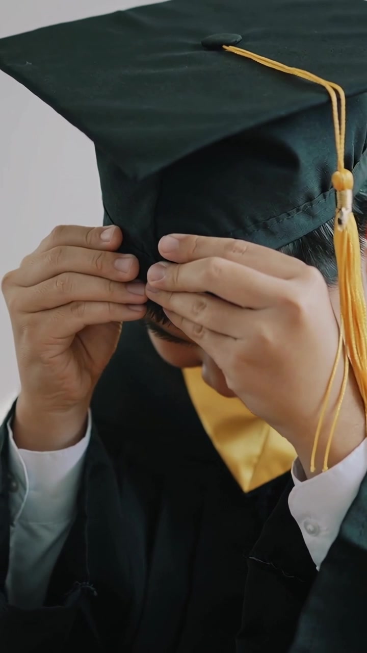 Video verticale: Laureato in toga che sistema il tocco in studio per la laurea, con stola e nappa dorata.