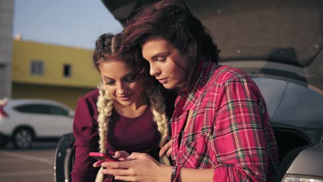 closeup view of two young attractive girlfriends looking at the smartphone, checking pictures while sitting inside of the open car trunk in the parking by the shopping mall during sunny day