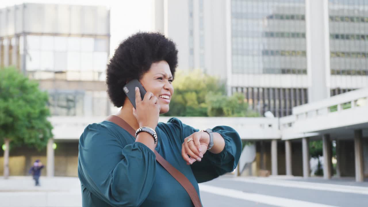 Happy plus size biracial woman talking on smartphone and checking smartwatch