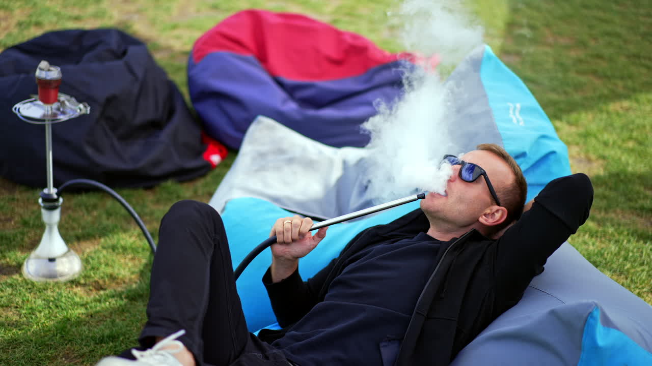 Having good rest outdoors smoking hookah. Mid-aged man in black casual clothes lies in bean bag chair and smokes shisha.
