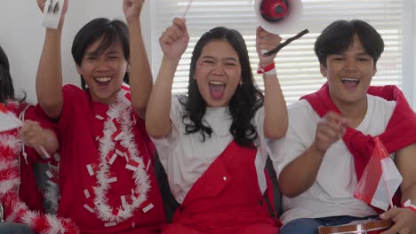 group of young man and woman indonesian supporters watching football match on tv and celebrating the goal using tambourine, megaphone and screaming