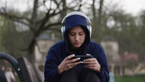 Mujer-Triste-Con-Capucha-Sentada-En-Un-Banco-Al-Aire-Libre,-Desplazándose-Por-El-Teléfono-Inteligente-Con-Auriculares-Puestos