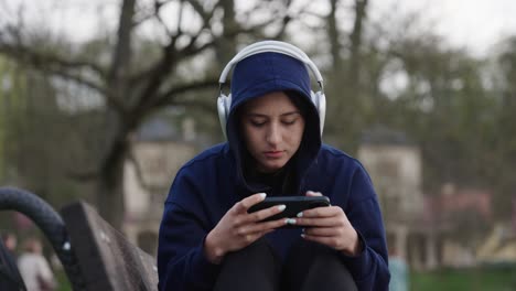 sad woman in hoodie sit on outdoor bench, scroll smartphone with headphones on