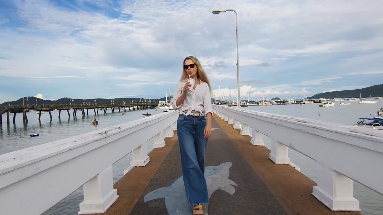 Woman walking on a pier by the ocean, drinking a beverage