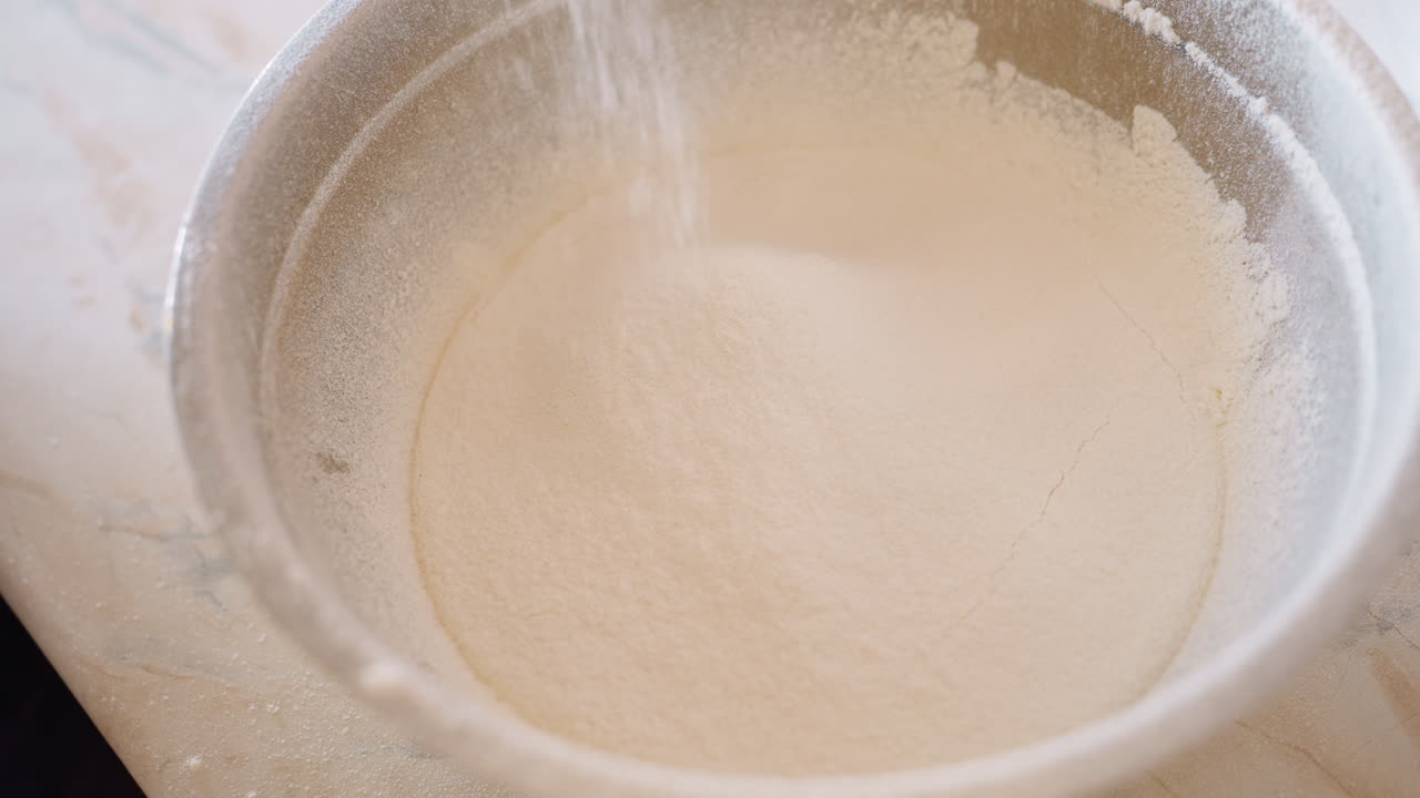 Fine flour being sifted into stainless bowl during baking preparation, smooth texture forming as ingredient settles evenly, creating perfect base for dough, bread, pastry or cake recipes