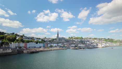view from a moving ferry or cruise ship as it passes the picturesque town of cobh, ireland. the scene includes the vibrant waterfront buildings, historic st. colman’s cathedral, and a clear sunny sky