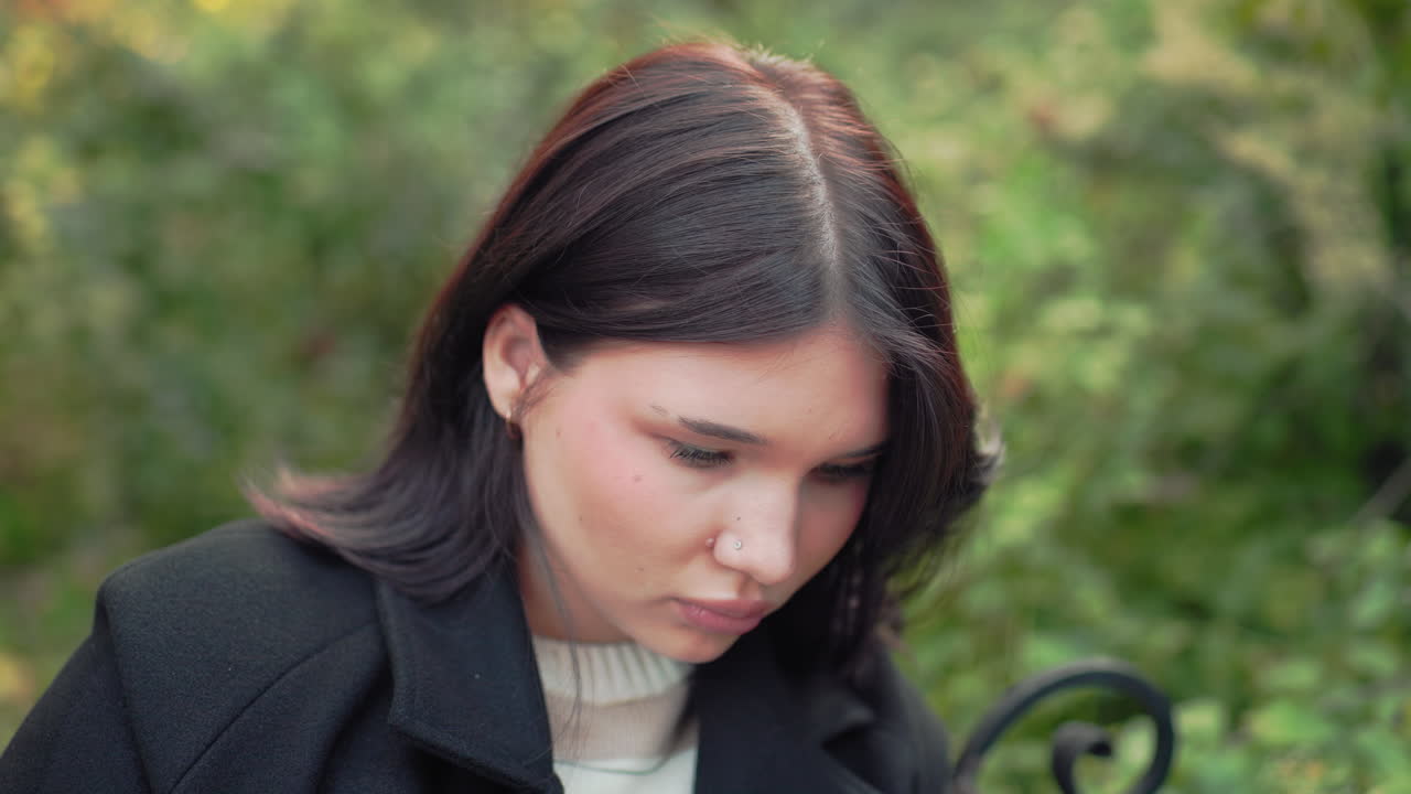 옆모습으로 코트와 스웨터를 입은 아늑한 분위기의 젊은 여성이 책 읽기에 집중하며, 공원 벤치에 앉아 머리카락을 귀 뒤로 넘기고 있어요. 부드러운 보케 배경에 은은한 가을의 녹음이 더해졌네요.