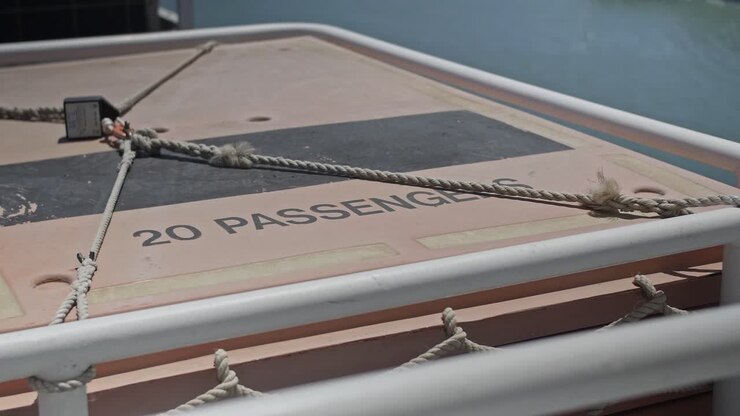 Una corda fissata che lega una zattera di salvataggio sul retro di un traghetto, con un cartello 'Max 20 passeggeri' in cima.