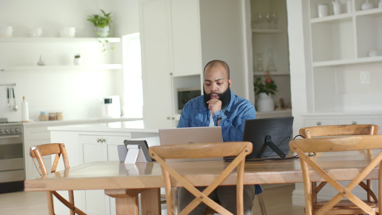Surrounded by devices, African American man studying main laptop and reviewing work in home kitchen
