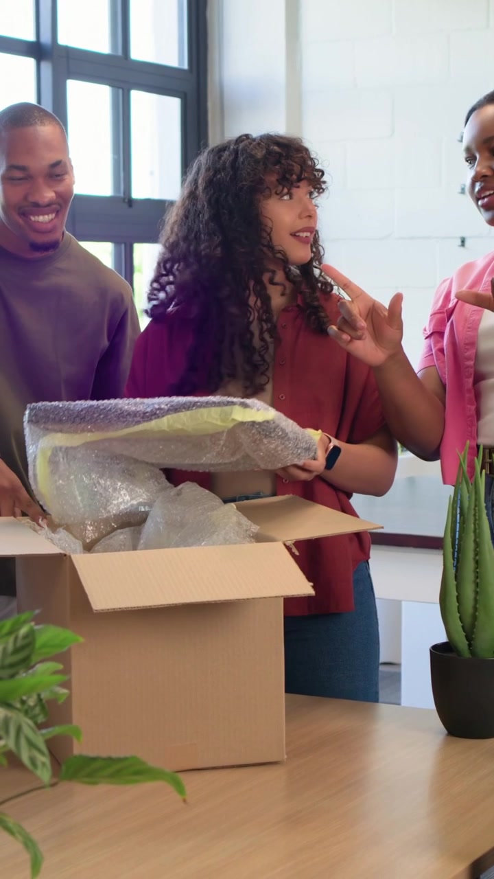 Vertical video: Opening box, woman in rust shirt lifting bubble-wrap print at table, team reviewing