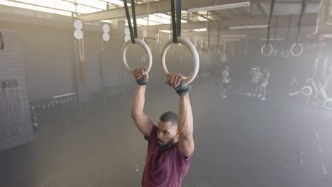 focused biracial man doing pull ups on gym rings at gym, in slow motion