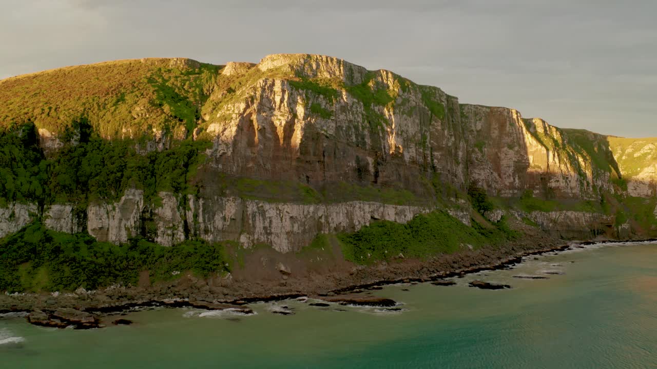 일몰 동안 뉴질랜드의 해안선을 따라 절벽을 때리는 태양 광선의 공중 궤도 샷