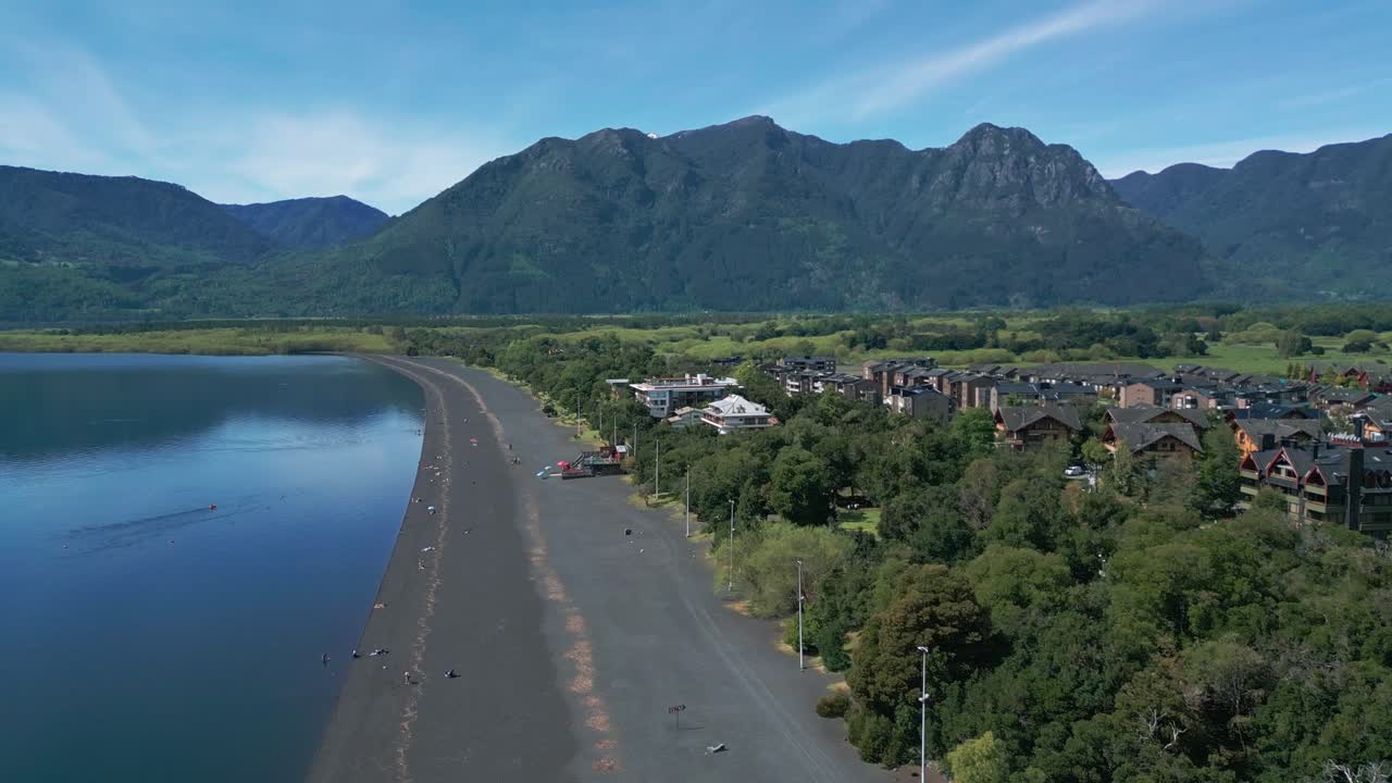 Aerial pan of a lakeside town in southern Chile with black sand beach, forest, and green mountains in the background