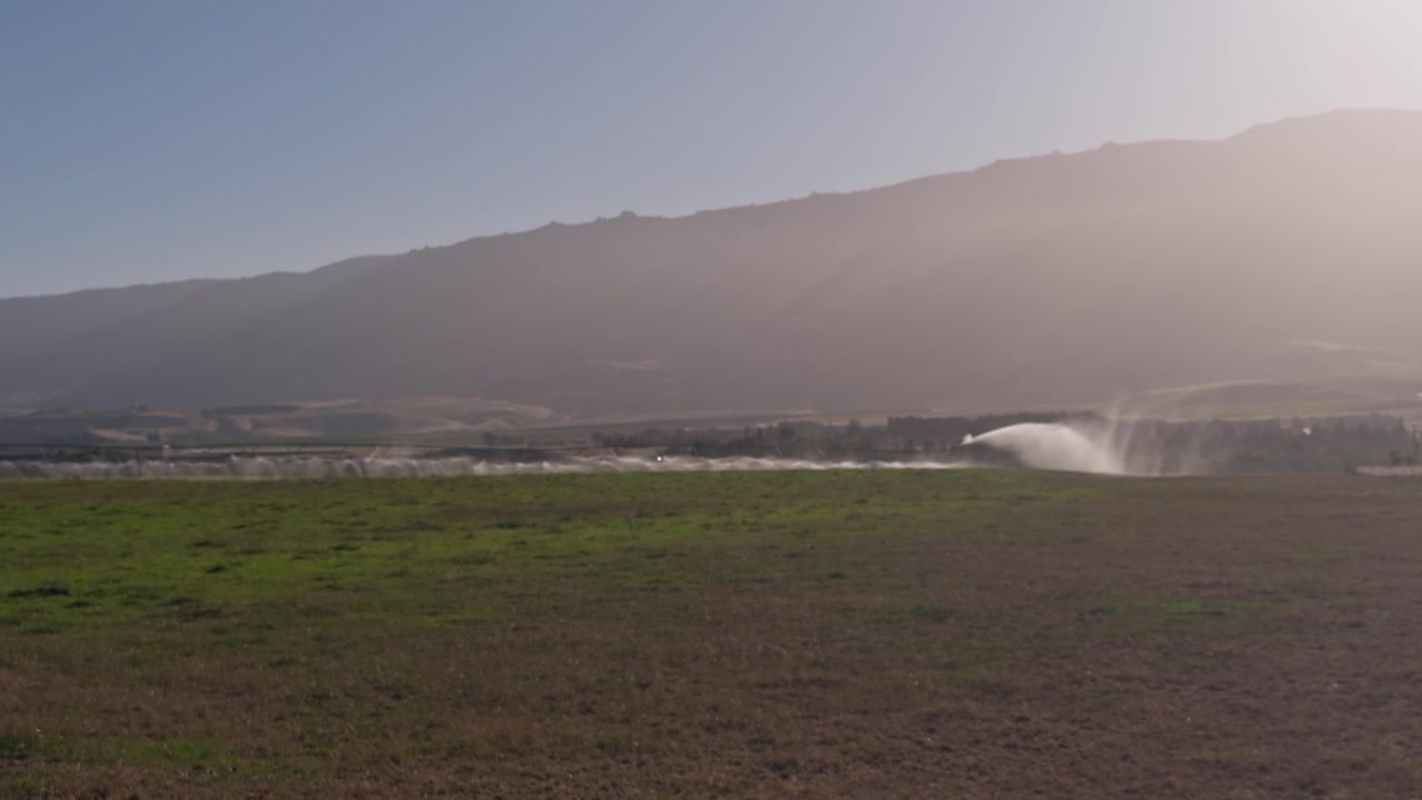 Un grande sistema di irrigazione a pivot centrale distribuisce l'acqua uniformemente su un campo agricolo. Terreno agricolo produttivo è circondato da dolci colline sotto un cielo azzurro brillante.