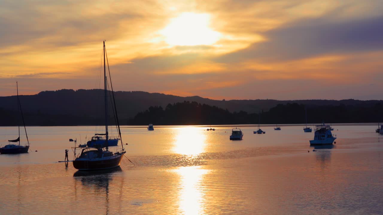 Un paddleboarder che passa accanto a barche ancorate durante il tramonto, lago Whakatane.