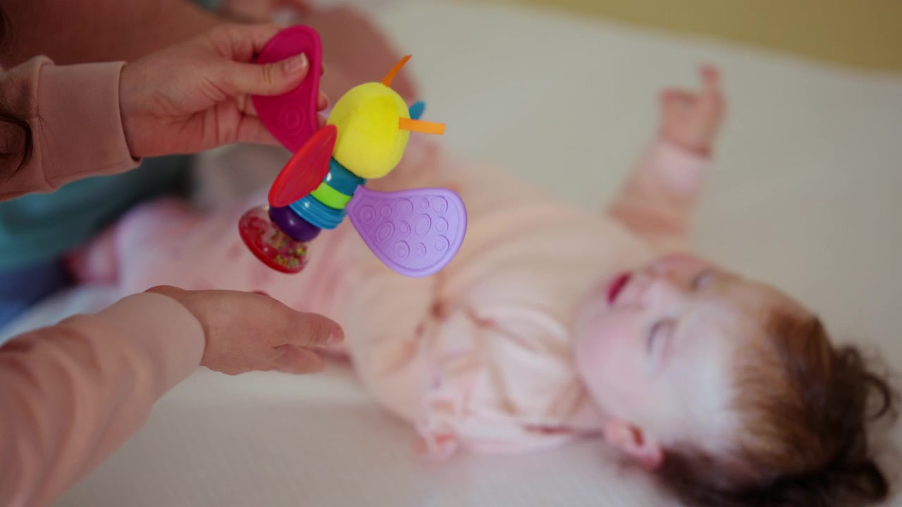 A colorful baby rattle is gently presented to an infant, while caregiver is performing mobility exercises