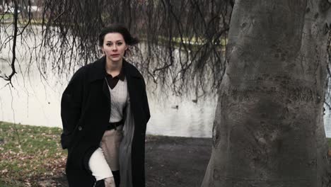 Junge-Frau-In-Einem-Schwarzen-Mantel,-Die-Im-Herbst-Am-Nachmittag-Unter-Einem-Windigen-Baum-In-Einem-Park-Steht-2