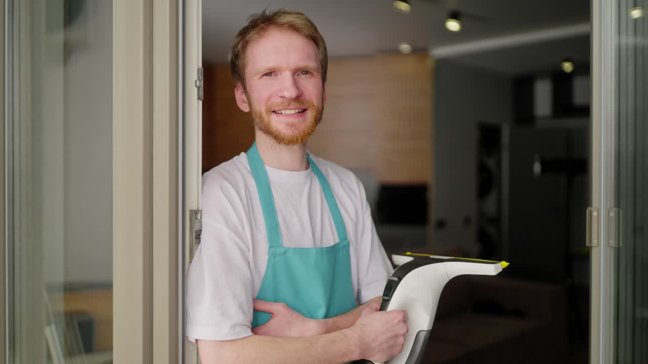 retrato de un hombre rubio confiado con una barba en una camiseta blanca y un delantal azul que sostiene una aspiradora en sus manos para limpiar ventanas en un apartamento moderno