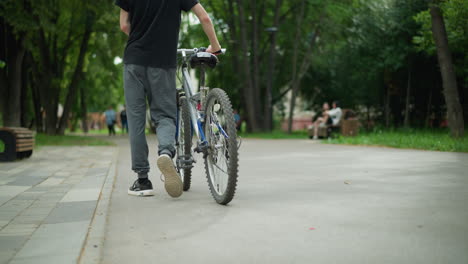 back view of young boy in black top and ash pants walking with his bicycle along park pathway, trees and greenery surround the scene, with blurred people sitting in the background