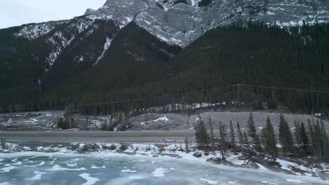 Aerial-view-pan-of-forested-frozen-lake-up-to-tall-snowy-mountains,-Alberta,-Canada---Drone-4k