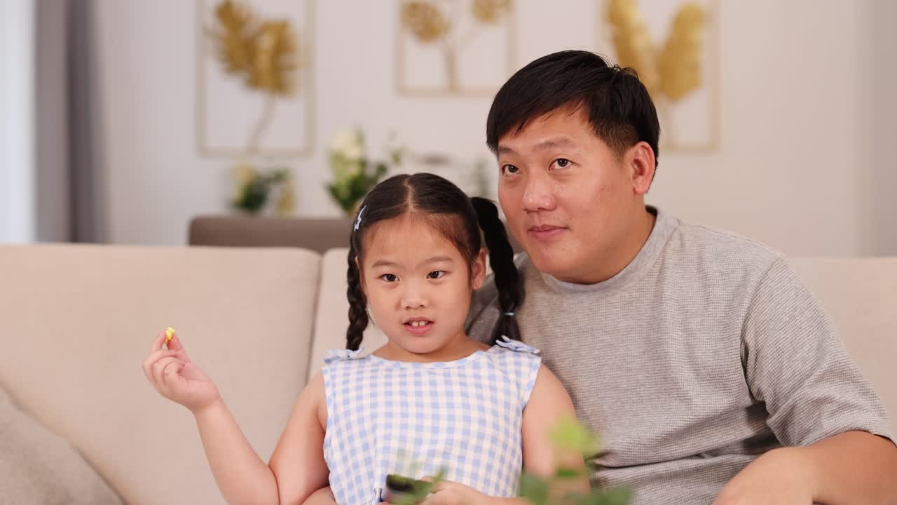 幸せそうなアジア系の父娘がソファでテレビを見ながらポップコーンを食べ、絆を深めている。