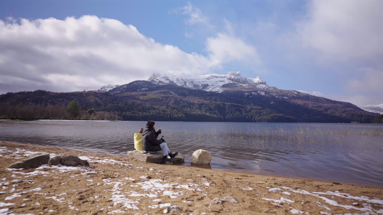 Parent with her kid sitting on snowy lake shore facing mountain while drinking mate