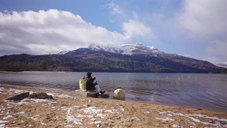 parent with her kid sitting on snowy lake shore facing mountain while drinking mate