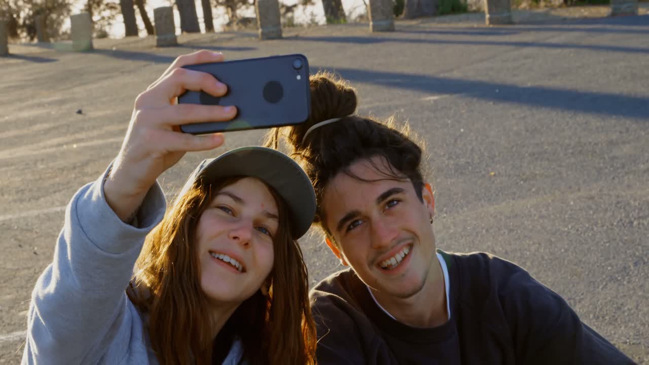 Vista frontale di una giovane coppia di skateboarder caucasici che si scatta un selfie con il cellulare su una strada di campagna 4K