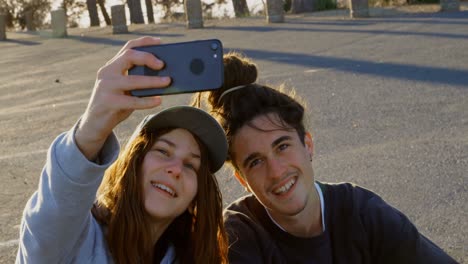 Vista-Frontal-De-Una-Joven-Pareja-De-Skaters-Caucásicos-Tomando-Selfie-Con-Un-Teléfono-Móvil-En-Una-Carretera-Rural-4k