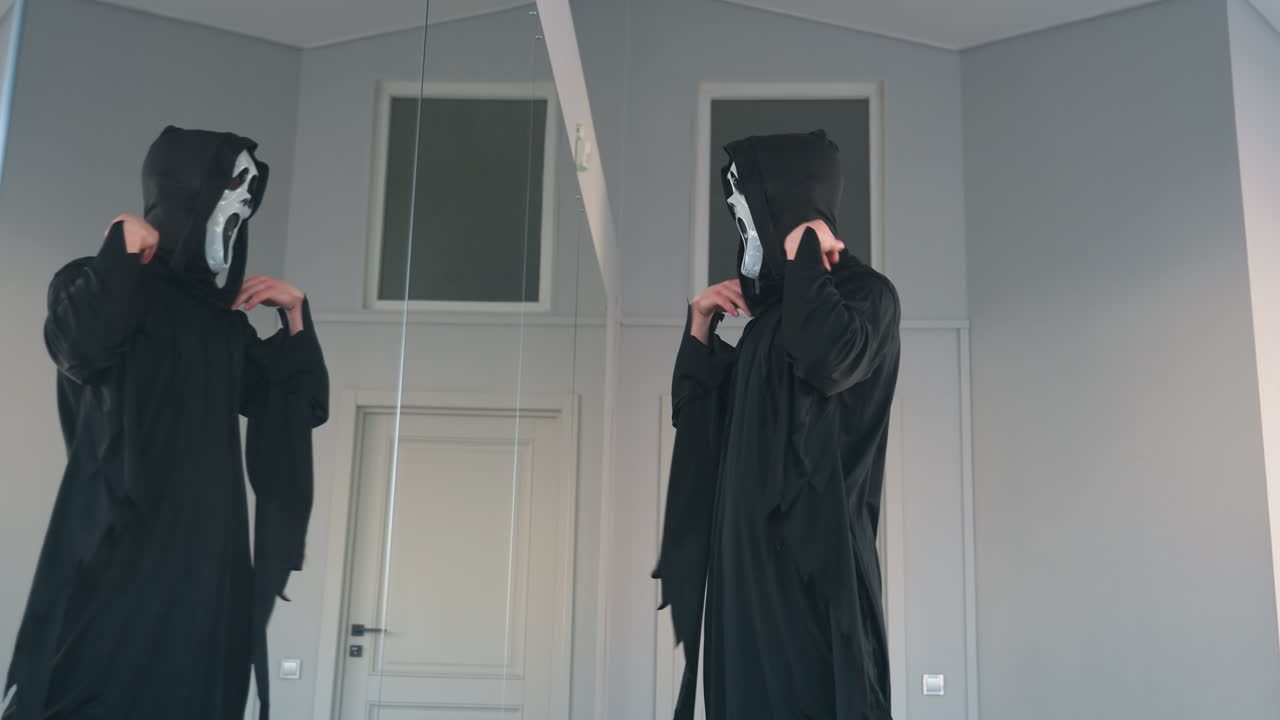 young man in dark tattered cloak stands before tall mirror, lifts ghostly white screaming mask to face, creating eerie silhouette against minimalist studio backdrop for Halloween transformation