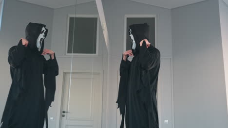 young man in dark tattered cloak stands before tall mirror, lifts ghostly white screaming mask to face, creating eerie silhouette against minimalist studio backdrop for halloween transformation