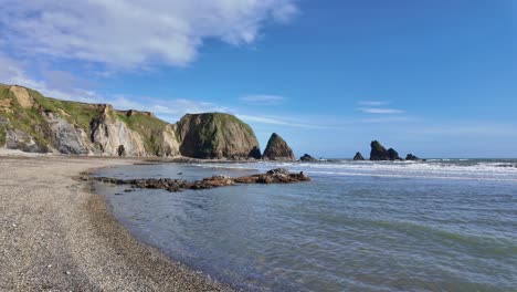 Kristallklares-Wasser,-Sanfte-Wellen-Am-Kieselstrand-Mit-Meeresklippen-Und-Blauem-Himmel-Mit-Bauschigen-Weißen-Wolken,-Strahlender-Frühlingstag,-Kupferküste,-Waterford,-Irland