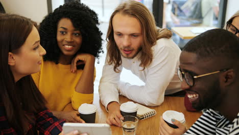 group multiethnic group of happy friends laughing and watching a video on a tablet while drinking coffe sitting at a table in a cafe