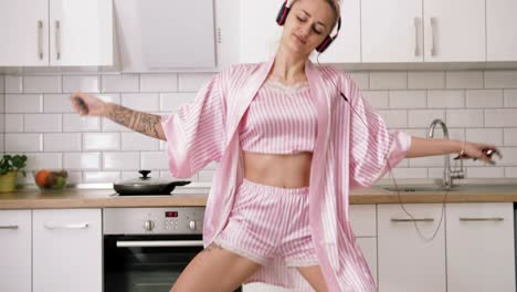 happy young woman with pink hair dancing in kitchen wearing pink pajamas and listening to music with headphones