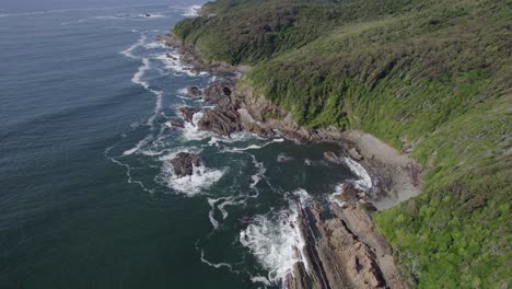 Foamy-Waves-Splashing-On-Rocky-Green-Cliffs-In-Forster,-Australia---aerial-drone-shot