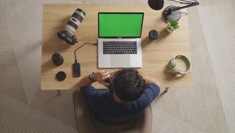 top view of a male video editor talking on smartphone while using a green screen laptop next to the camera in the workspace at home