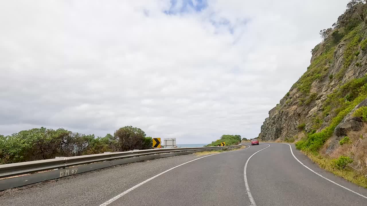 A car travels along the winding Great Ocean Road, surrounded by coastal cliffs and ocean views under a cloudy sky