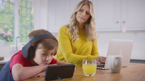 boy in kitchen watching movie on mobile phone wearing wireless headphones as mother works on laptop