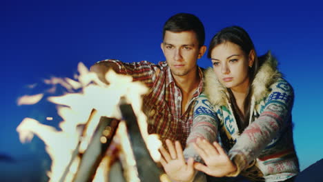 A-Loving-Young-Couple-Sitting-On-The-Beach-Campfire-Warm-Hands-By-The-Fire-Romance-And-Youth