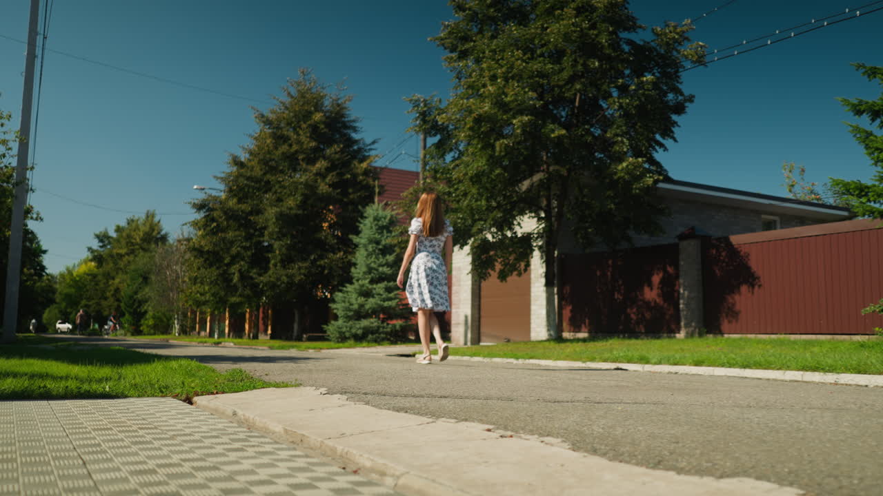 Lady walks confidently on sunlit paved street bordered by greenery and residential buildings while distant figures include cyclist and other pedestrians moving in same direction under clear blue sky
