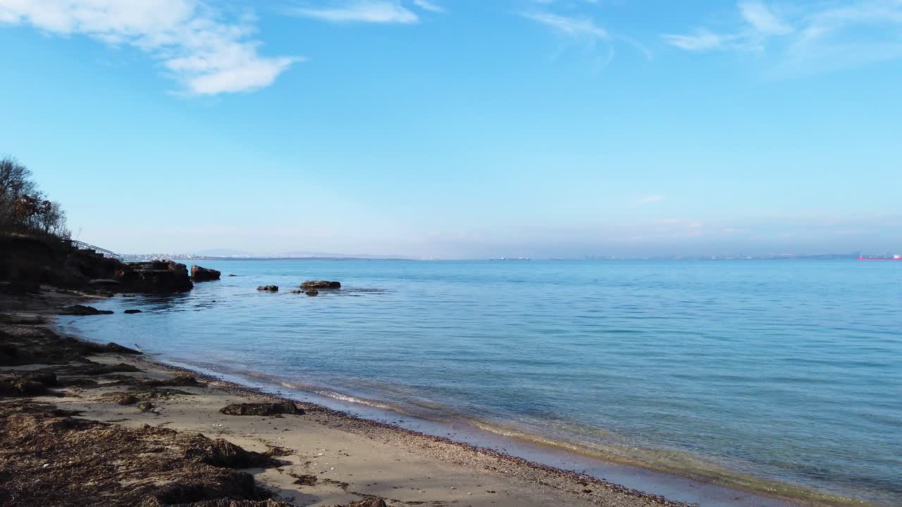 Sea view from the beach after sunrise. Calm sea, rocks in the sea water, algae on the shore. Blue sky, with a slight pink tinge. Trees by the shore. Winter morning to Black sea. Pan right