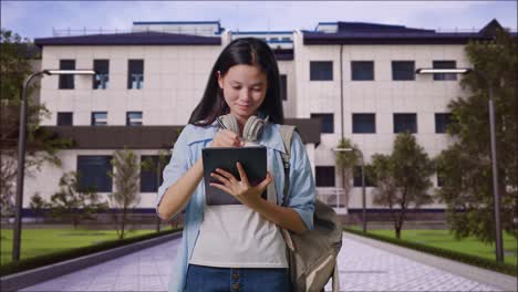 estudiante adolescente asiática con una mochila sonriendo y tomando nota en la tableta mientras está de pie frente a un edificio escolar