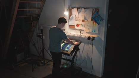 man uses touchscreen control console in dark workspace under staircase, surrounded by cables, notes, lighting charts, and documents on bulletin board, adjusting settings for studio production or video operation