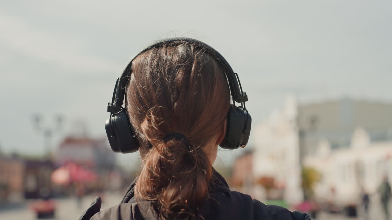 Jovem com fones de ouvido, de costas para a câmera em uma praça ensolarada, pedestres desfocados no plano de fundo, rabo de cavalo solto, jaqueta escura, caminhada lenta, clima contemplativo, tráfego ambiente, cinematográfico