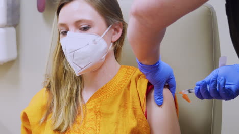 COVID-19-Vaccine-Shot-in-the-Arm-of-Young-Adult-Female-Woman-Wearing-a-Medical-Mask,-Corona-Virus-Injection-Administered-by-Nurse-in-Shoulder-in-Doctor’s-Office-with-the-Patient-Person’s-Face-Visible