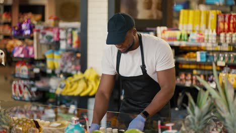 A-man-with-Black-skin-in-a-white-T-shirt-and-a-Black-apron-using-blue-gloves-manipulates-goods-on-the-counter-in-a-large-grocery-store-and-his-employee-a-man-in-a-gray-T-shirt,-clarifies-questions-and-consults-with-him